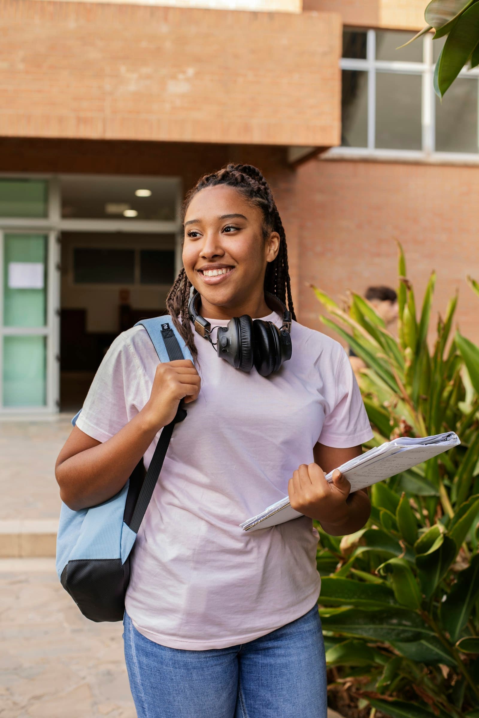 A smiling student with a backpack and notebooks, outside a campus building