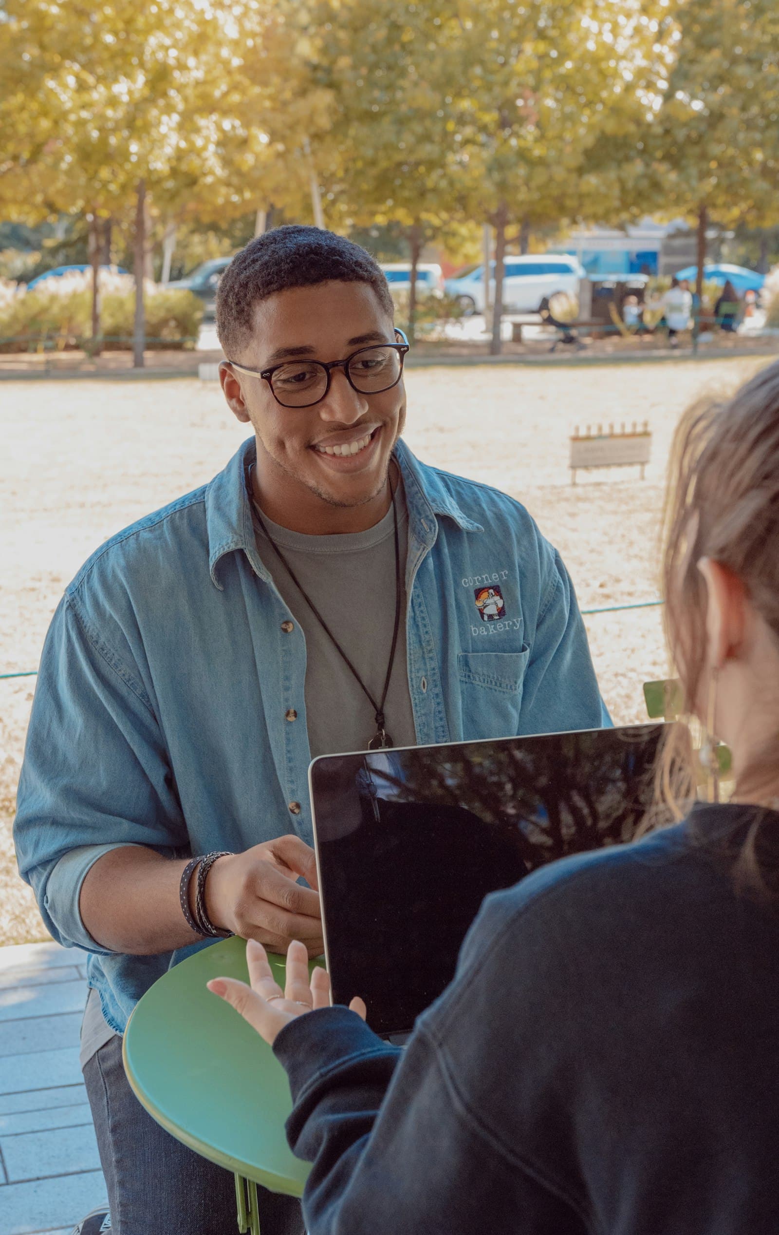 A smiling member of staff wearing a lanyard, talking with a student across a laptop on a campus terrace